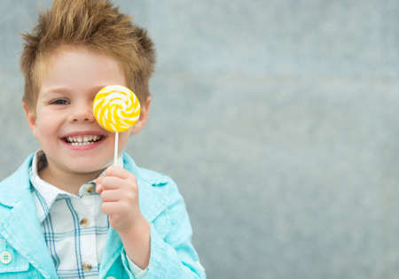 Fashion baby boy in mint jacket and sneakers stands on a gray wall background. Trendy boy with lollipop standing on the street.の写真素材