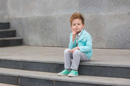 Fashion baby boy in mint jacket and sneakers posing on a gray wall background. Trendy boy standing...