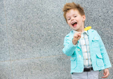 Fashion baby boy in mint jacket and sneakers stands on a gray wall background. Trendy boy with lollipop standing on the street.の写真素材