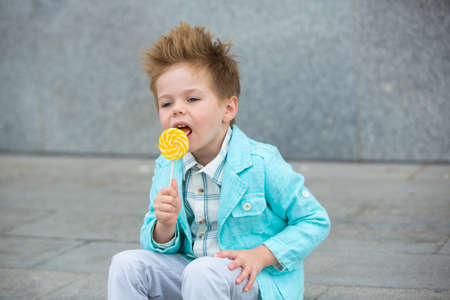 Fashion baby boy in mint jacket and sneakers stands on a gray wall background. Trendy boy with lollipop standing on the street.の写真素材