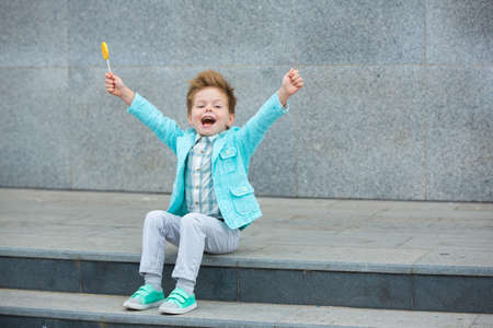 Fashion baby boy in mint jacket and sneakers stands on a gray wall background. Trendy boy with lollipop standing on the street.の写真素材