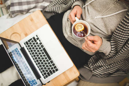 Young man laying in bad with laptop and cup of tea in hand.の写真素材