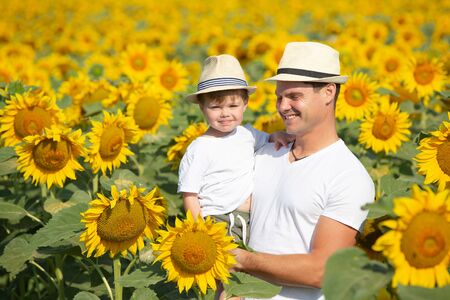 A child and his dad are playing in a huge field of sunflowers. Little boy and man in white t-shirts and hats. Dad holds the baby in his armsの写真素材