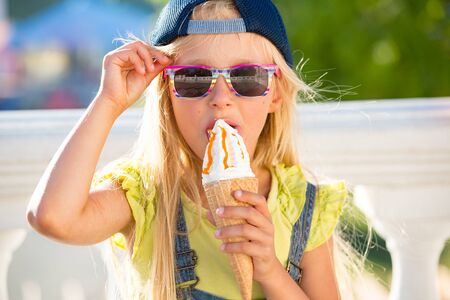 Little girl enjoy ice cream in a vaiel glass. Teen is fashionably dressed. Child posing against the backdrop of street sceneryの写真素材