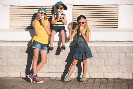 Two sisters and their brother enjoy ice cream in a vaiel glass. Teenagers are fashionably dressed. Children posing against the backdrop of street sceneryの写真素材