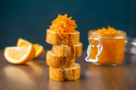 A jar of orange confiture and pieces of French baguette with star anise and sliced orange fruit on ...