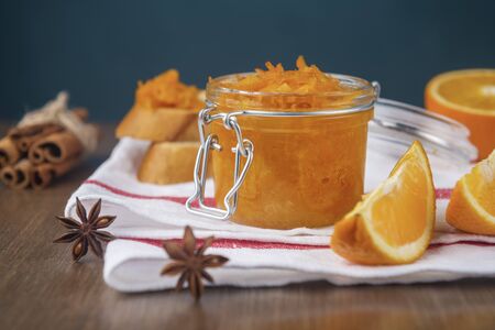 Jar of orange jam on a white napkin with slices of French baguette, sliced orange, cinnamon and star anise on a wooden tableの写真素材