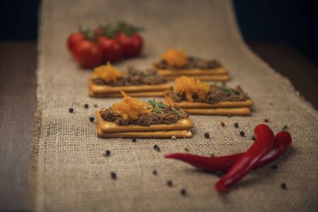 A few crackers with poultry pate and orange jam, red cocktail tomatoes and hot pepper on the sackcloth and wooden background, homemade liver paste decorated with spices on the burlap wooden tableの写真素材