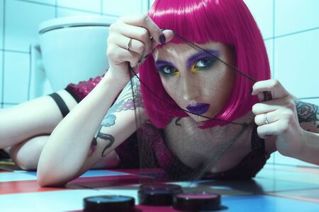 Beautiful girl in red dress with purple lips and pink hair lying in the bathroom on the colored tiles floorの写真素材