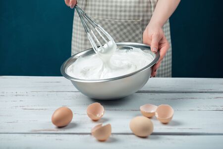 Woman in checkered apron whips egg whites on a white vintage wooden table. The process of making meringues.の写真素材