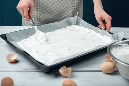 Woman spreads beaten egg whites on a baking sheet covered with parchment on a white vintage wooden table. The process of making meringues.の写真素材