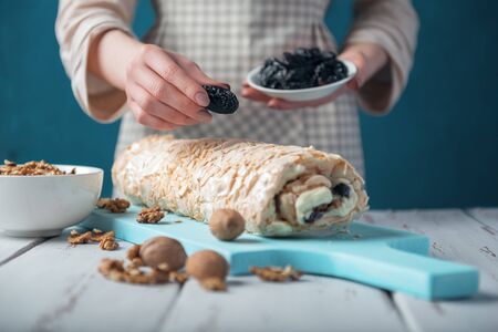 Woman in checkered apron decorating meringue roll with prunes on a white vintage wooden kitchen table. The process of making meringue roll. Meringue pie and bowls with prunes and walnuts.の写真素材