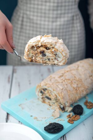 Woman serves with cake server a piece of meringue roll on a white vintage wooden kitchen table. Meringue pie decorated with prunes and walnuts on blue cutting board.の写真素材