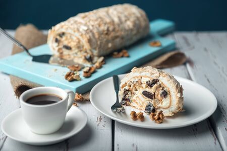 A cup of coffee and meringue roll on a white vintage wooden kitchen table with burlap napkin. Meringue pie decorated with prunes and walnuts on blue cutting board.の写真素材