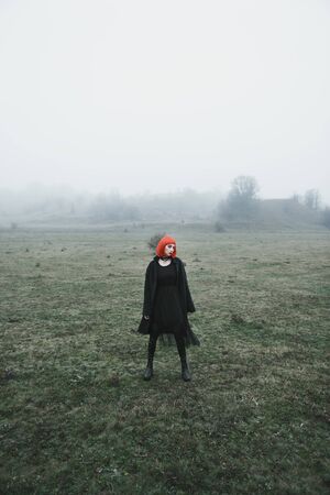 Fashion girl with red bob haircut and black clothes in dramatic cold foggy autumn fieldの写真素材