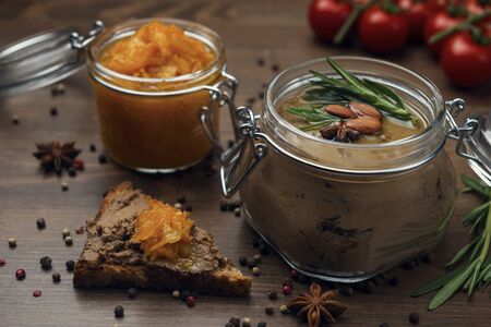 A slice of bread with poultry pate and orange gam, glass jar with poultry pate, decorated with almonds and rosemary, and jar with orange jam on a wooden table. Homemade liver paste decorated with spicesの写真素材
