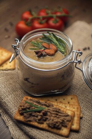 Glass jar with poultry pate, decorated with almonds and rosemary, crackers with pate and red cocktail tomatoes on the sackcloth and wooden background, homemade liver paste decorated with spices on the burlap wooden tableの写真素材