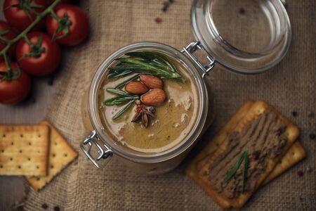 Glass jar with poultry pate, decorated with almonds and rosemary, crackers with pate and red cocktail tomatoes on the sackcloth and wooden background, homemade liver paste decorated with spices on the burlap wooden tableの写真素材