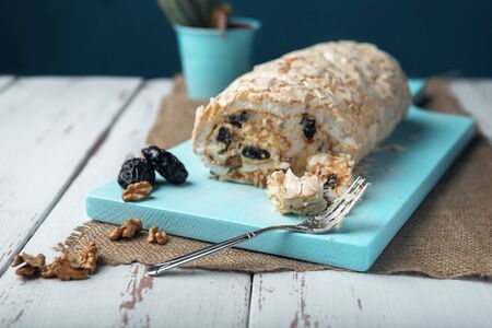 A piece of meringue roll and meringue roll on a white vintage wooden kitchen table with burlap napkin. Meringue pie decorated with prunes and walnuts on blue cutting board.の写真素材