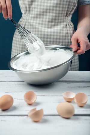 Woman in checkered apron whips egg whites on a white vintage wooden table. The process of making meringues.の写真素材