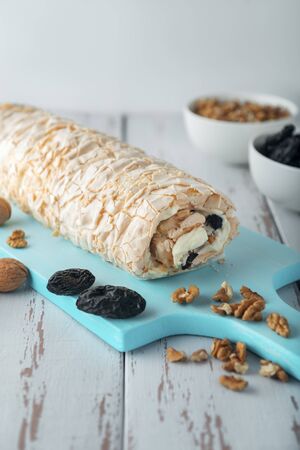 Prune-nut meringue roll on a blue cutting board on a white vintage wooden kitchen table. Meringue pie and bowls with prunes and walnuts.の写真素材