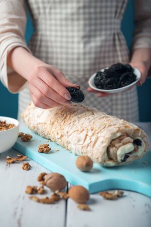 Woman in checkered apron decorating meringue roll with prunes on a white vintage wooden kitchen table. The process of making meringue roll. Meringue pie and bowls with prunes and walnuts.の写真素材
