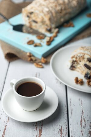 A cup of coffee and meringue roll on a white vintage wooden kitchen table with burlap napkin. Meringue pie decorated with prunes and walnuts on blue cutting board.の写真素材