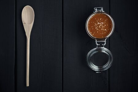 Glass jar of salted caramel with a wooden spoon on a black wooden table. Top view.の写真素材