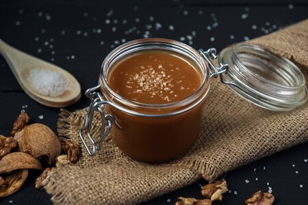 Glass jar of salted caramel with a wooden spoon of salt, walnuts and nutshells on a black wooden table with burlap napkinの写真素材