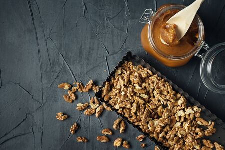 Glass jar of salted caramel with wooden spoon and walnuts on dark background. Flat lay of preparation for cooking tart. Space for text.の写真素材