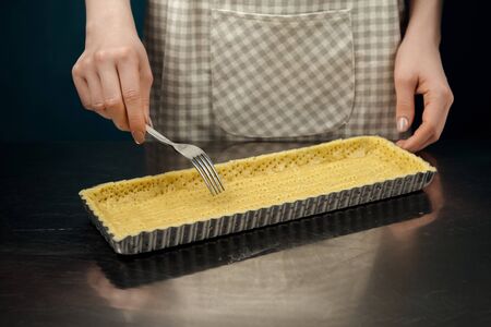Woman in checkered apron piercing the dough with a fork for blind baking. Woman's hands making cookie pastry for tart.の写真素材