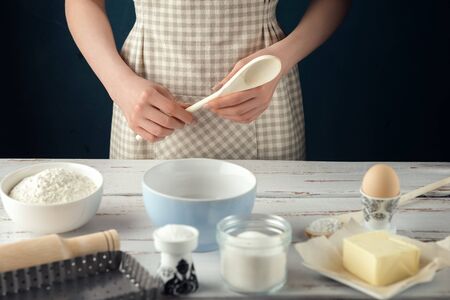 Woman in checkered apron with wooden spoon behind kitchen table. Ingredients and baking utensils for cooking tart, cookies, pastry: egg, flour, butter, rolling pin, sugar, salt の写真素材