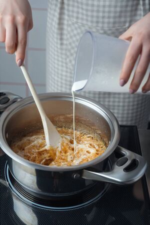 Woman in a checkered apron places cream in a stainless steel pan while making caramelの写真素材