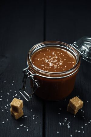 A glass jar with homemade salted caramel and a few pieces of brown sugar with salt crystals on the black backgroundの写真素材