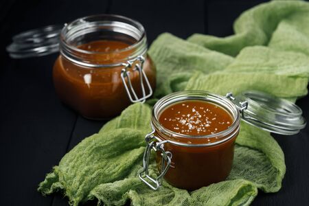 Two glass jars of homemade caramel with salt crystals on green gauze and black wooden tableの写真素材