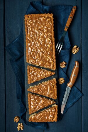 Chopped walnut-caramel tart on a black serving board with walnuts and cutlery on dark wooden table. Top view. Space for text.の写真素材