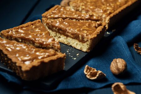 Chopped walnut-caramel tart on a black serving board with walnuts on dark wooden tableの写真素材