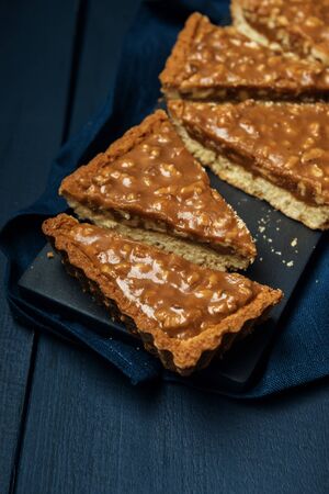 Chopped walnut-caramel tart on a black serving board on dark wooden tableの写真素材