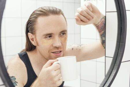 Unshaven man with patches under his eyes and cup of coffee looks at himself in the bathroom mirror.の写真素材