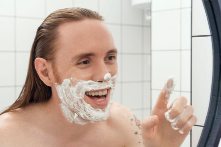 Tattooed man with long hair with shaving foam on his face smiling in front of the bathroom mirror.