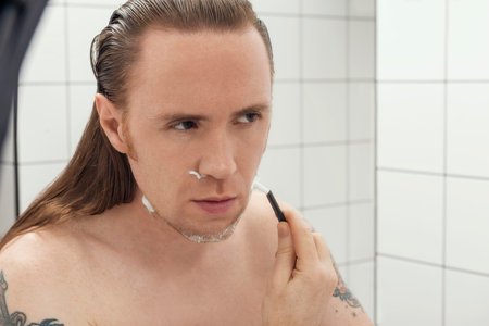 Tattooed man with long hair shaving his face in front of the bathroom mirror.