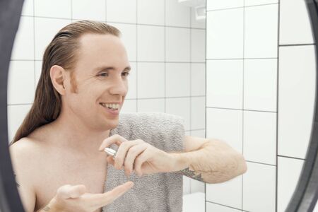 A man with tattoos and long hair sprays on his hand after-shave lotion in the bathroomの写真素材