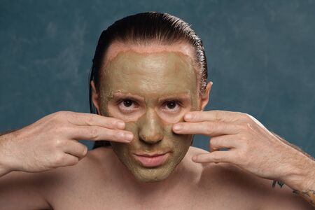 A tattooed man with long wet hair is applying a green clay mask with his hands.の写真素材