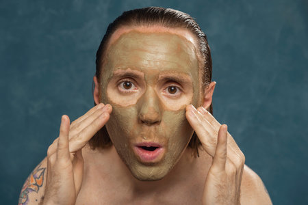 A man with long wet hair is applying a green clay mask with his hands. Male spa procedures.