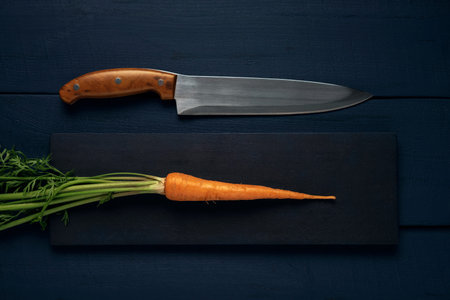 One single raw carrot with green leaves and kitchen knife on a wooden table. Top view.