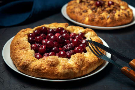 Two galettes with ripe red cherry and walnut filling on dark blue background. Homemade sweet open pie with napkin and cutlery. Close-up.の写真素材