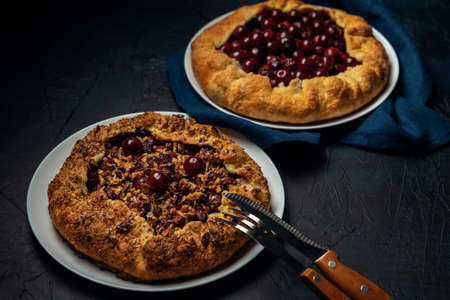 Two galettes with ripe red cherry and walnut filling on dark blue background. Homemade sweet open pie with napkin and cutlery. Close-up.の写真素材