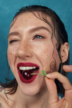 Close-up portrait with wide angle of funny freckled girl with cherry berry. Funny fashion girl bites cherry in front of the blue background.の写真素材