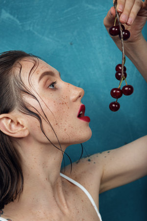 Young fashion model with freckles look at cherry berries in front of the blue background.