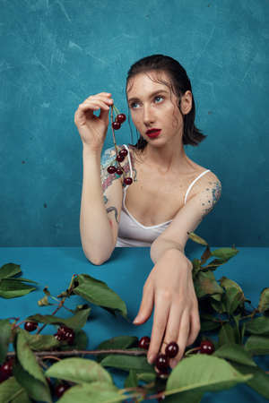 Dreamed female fashion model sitting at the table with cherry branch in front of the blue backgroundの写真素材